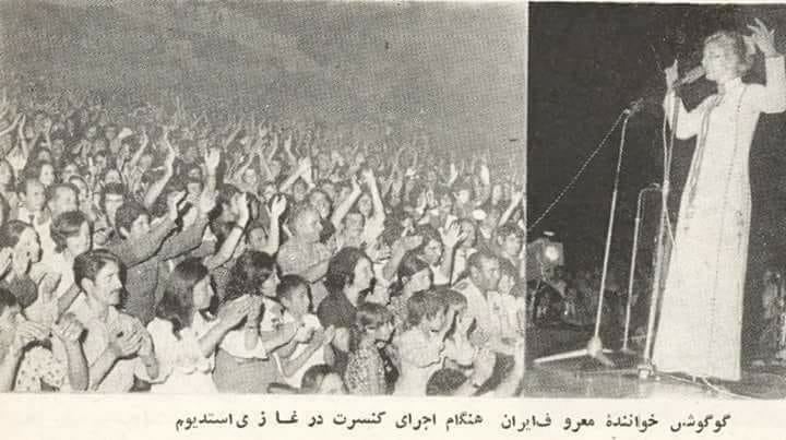 A black-and-white photograph from the 1970s showing Googoosh, an Iranian singer, performing on stage at Ghazi Stadium in Kabul, Afghanistan, with an audience of diverse men, women, and children clapping in appreciation.